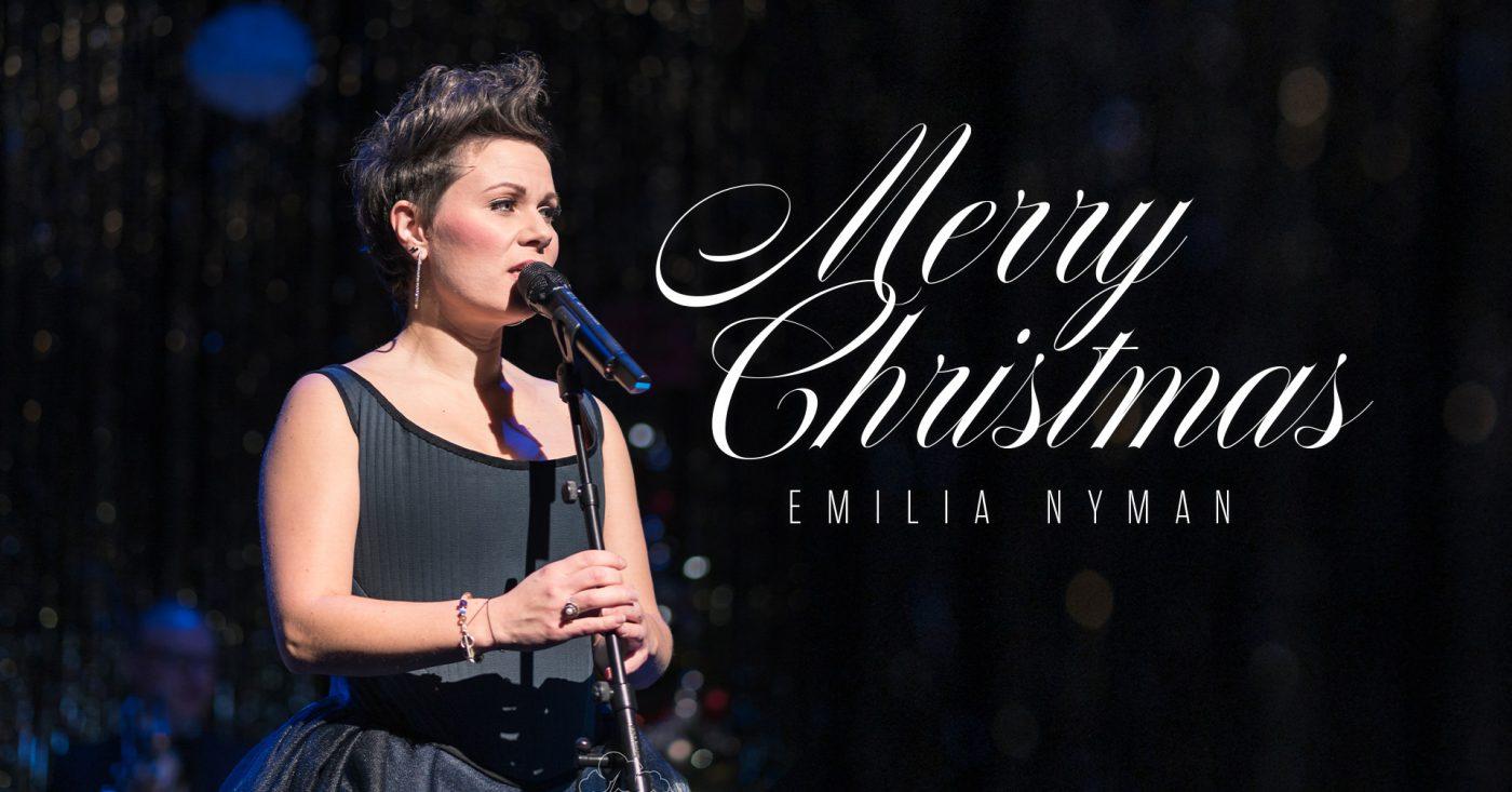 Kuvassa Merry Christmas konsertin laulaja Emilia Nyman mikrofoni kädessä tummaa taustaa vasten.
